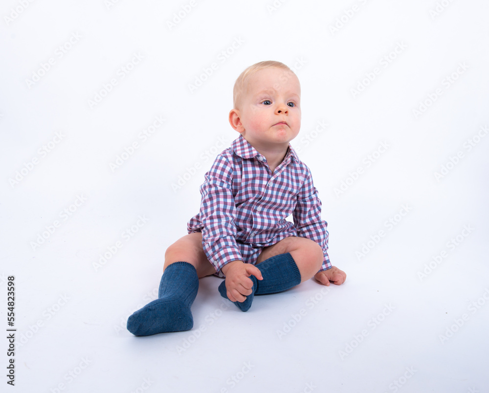 Playful beautiful baby boy wearing plaid bodysuit standing over white studio background sitting and smiling to the camera. Baby boy wants to play with his mother Playful beautiful baby boy wearing plaid bodysuit standing over white studio background sitting and smiling to the camera. Baby boy wants to play with his mother
