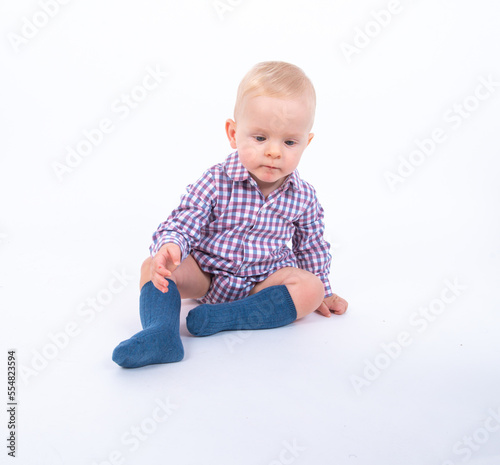 Playful beautiful baby boy wearing plaid bodysuit standing over white studio background sitting and smiling to the camera. Baby boy wants to play with his mother 