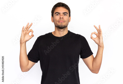 Calm young man with mudra gesture. Portrait of thoughtful Caucasian male model with short dark hair in black T-shirt looking at camera with raised hands, practicing meditation. Yoga concept