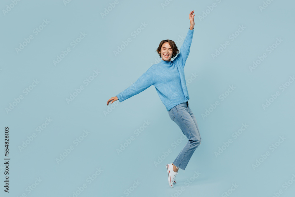 Full body young woman wear knitted sweater look camera stand on toes leaning back with outstretched hands isolated on plain pastel light blue cyan background studio portrait. People lifestyle concept.