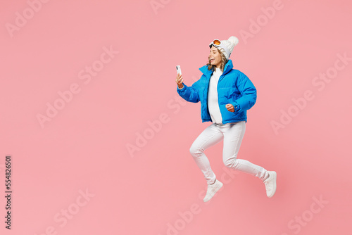 Snowboarder fun woman wear blue suit goggles mask hat ski padded jacket jump hold use mobile cell phone isolated on plain pastel pink background. Winter extreme sport hobby weekend trip relax concept.
