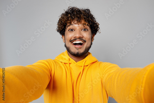 Close up young cheerful happy fun Indian man 20s he wearing casual yellow hoody doing selfie shot pov on mobile cell phone isolated on plain grey background studio portrait. People lifestyle portrait.