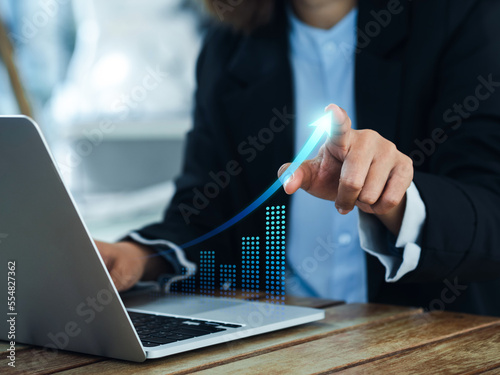 Investment technology, financial, return on investment - ROI concepts. Increasing arrow, the exponential curve of progress in business performance over charts on laptop draws by businesswoman's hand. photo