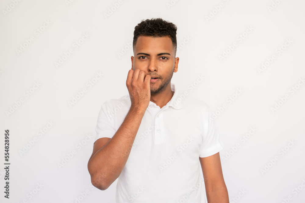 Stressed young man biting his fingernail. Male Indian model with brown eyes and curly hair in white polo shirt looking bored hearing something uninteresting. Boredom, dissatisfaction concept Stressed young man biting his fingernail. Male Indian model with brown eyes and curly hair in white polo shirt looking bored hearing something uninteresting. Boredom, dissatisfaction concept