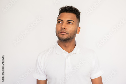 Portrait of pensive young man looking away. Male Indian model with brown eyes and curly hair in white polo shirt dreaming or making important decision. Thought, dream concept