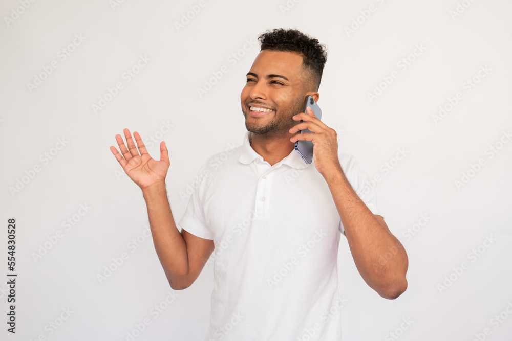 Cheerful young man talking on mobile phone waving his hand. Male Indian model with brown eyes and curly hair in casual clothes smiling speaking on phone. Communication, modern technology concept Cheerful young man talking on mobile phone waving his hand. Male Indian model with brown eyes and curly hair in casual clothes smiling speaking on phone. Communication, modern technology concept