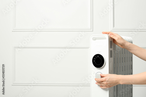 Young woman adjusting temperature on modern electric heater near white wall, closeup. Space for text