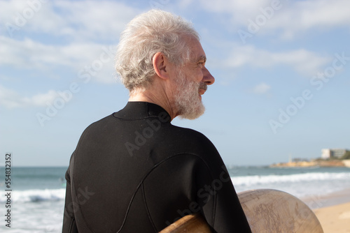 Portrait of sporty aged man with surfboard. Caucasian grey-haired man walking along seaside holding board going to surf, relaxed and looking far. Sport and healthy lifestyle of mature people concept