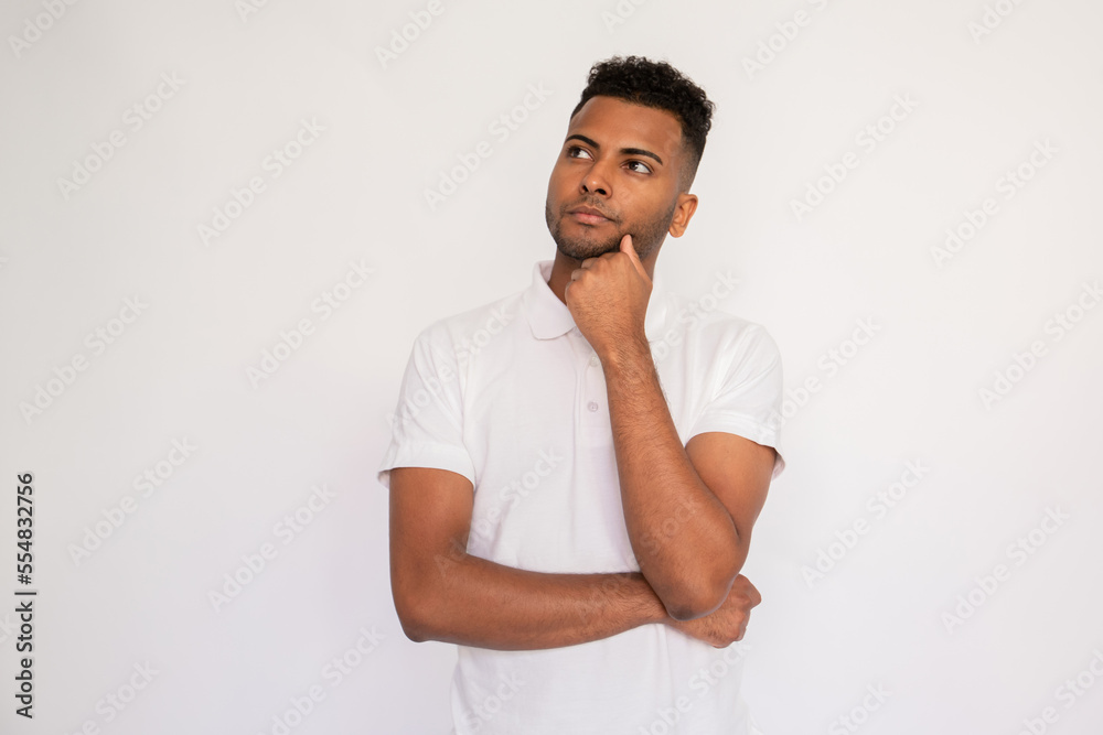 Portrait of pensive young man standing with hand on chin over white background. Latin American guy wearing white T-shirt looking away with thoughtful expression. Thinking and planning concept Portrait of pensive young man standing with hand on chin over white background. Latin American guy wearing white T-shirt looking away with thoughtful expression. Thinking and planning concept