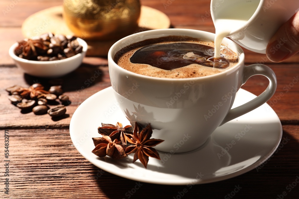 Woman pouring milk into cup of coffee with anise stars at wooden table