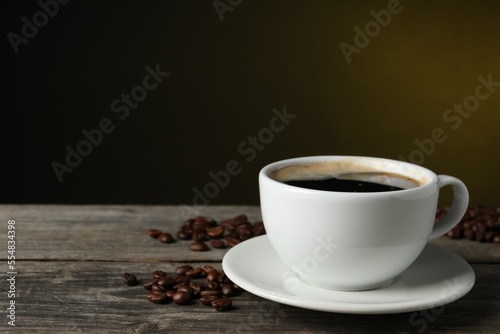 Cup of hot aromatic coffee and roasted beans on wooden table against dark background, space for text