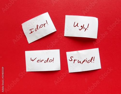 Torn paper pieces on red background with bully words UGLY STUPID WEIRDO IDIOT, concept of someone who hurts or frightens victims by degrade or demean in some way to feel powerful or stronger