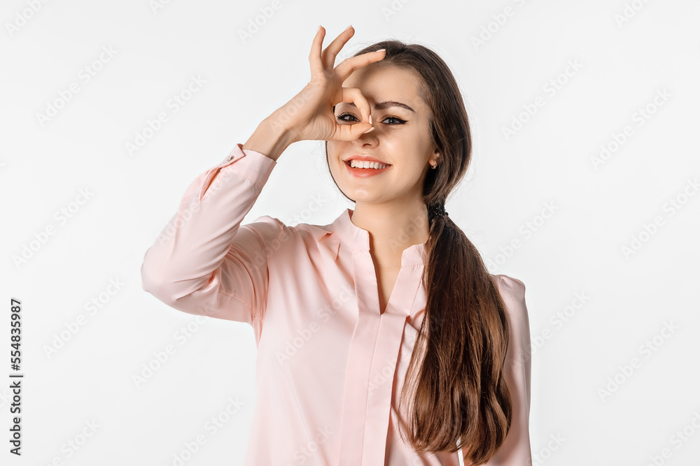 Excited young brunette woman is looking at camera, showing Ok sign and smiled, eye looking through fingers with happy face, standing against white studio background Excited young brunette woman is looking at camera, showing Ok sign and smiled, eye looking through fingers with happy face, standing against white studio background