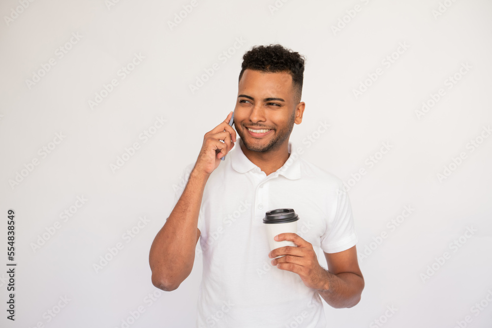 Office employee talking on mobile phone holding cup in his hand. Male Indian model with brown eyes and curly hair in white polo shirt speaking on phone holding cup of beverage. Coffee break concept Office employee talking on mobile phone holding cup in his hand. Male Indian model with brown eyes and curly hair in white polo shirt speaking on phone holding cup of beverage. Coffee break concept