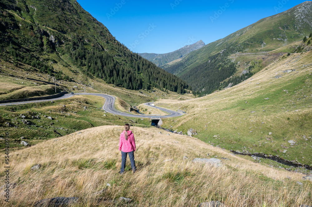 Rear view of a young woman standing at the famous picturesque curvy road in Fagaras mountain Romania. Beautiful girl enjoying the beauty of nature.