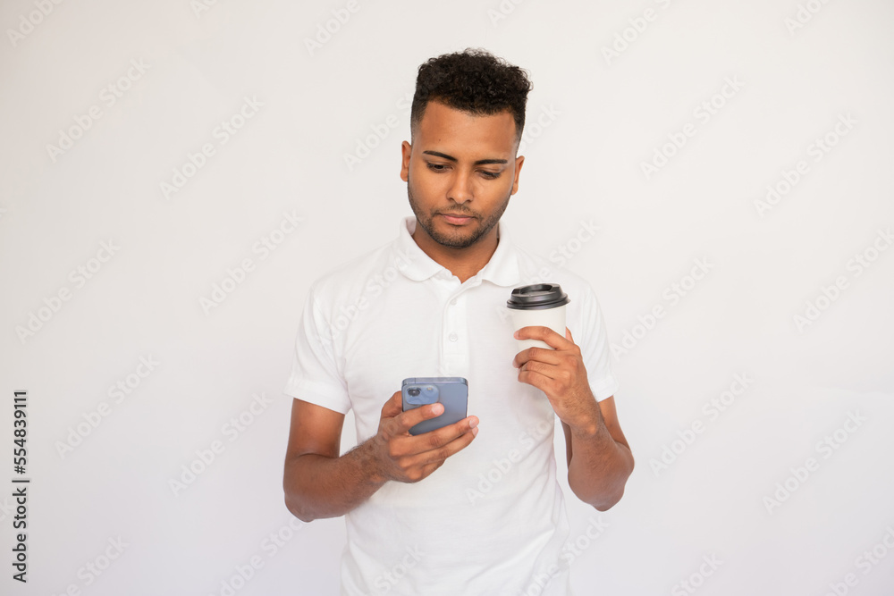 Office employee with smartphone and cup in his hands. Male Indian model with brown eyes and curly hair in white polo shirt using smartphone and holding cup of beverage. Coffee break concept Office employee with smartphone and cup in his hands. Male Indian model with brown eyes and curly hair in white polo shirt using smartphone and holding cup of beverage. Coffee break concept
