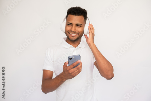 Joyful young man listening to music using his smartphone.Male Indian model with brown eyes and curly hair in white polo shirt wearing headphones enjoying music.Modern technology, advertisement concept © KAMPUS