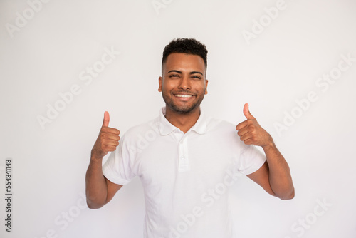 Portrait of happy young man making thumbs up over white background. Latin American guy wearing white T-shirt showing like gesture and smiling at camera. Approval concept © KAMPUS