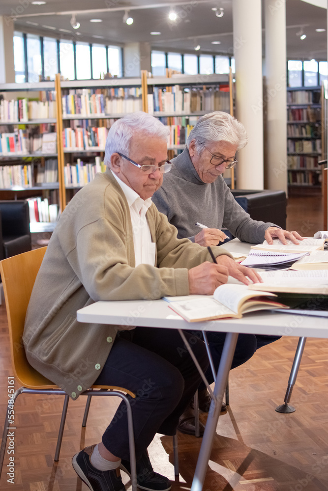 Side view of serious aged men studying with joy. Two grey-haired men concentrated on preparing for computer courses reading books and writing down necessary information. Adult people education concept Side view of serious aged men studying with joy. Two grey-haired men concentrated on preparing for computer courses reading books and writing down necessary information. Adult people education concept