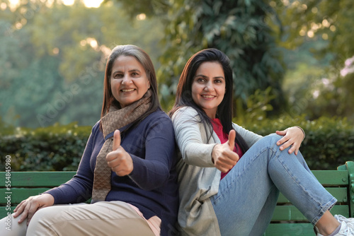 Two indian woman sitting at park in winter-wear and showing thumps up.