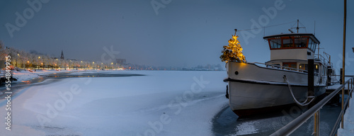 Ferry with Christmas tree standing at edge of bow. Panorama of ship boat moored at the pier covered with snow and promenade in winter in Schleswig. Winter, Christmas time on the Schlei, Schleswig. photo