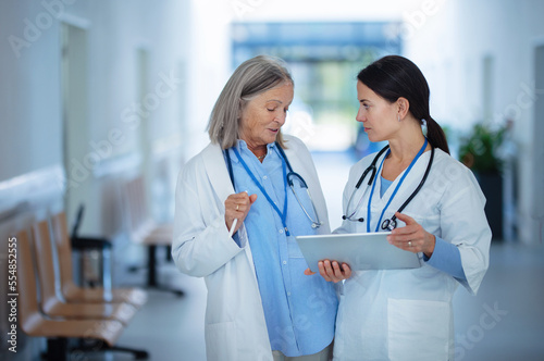 Fotografie Older doctor giving advise to her younger colleague, discussing at hospital corridor