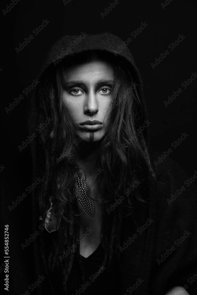 Beautiful woman studio portrait. Model with make-up, dreadlocks, feather attached to her hair hiding under hood and looking to camera with seductive look. Black and white image