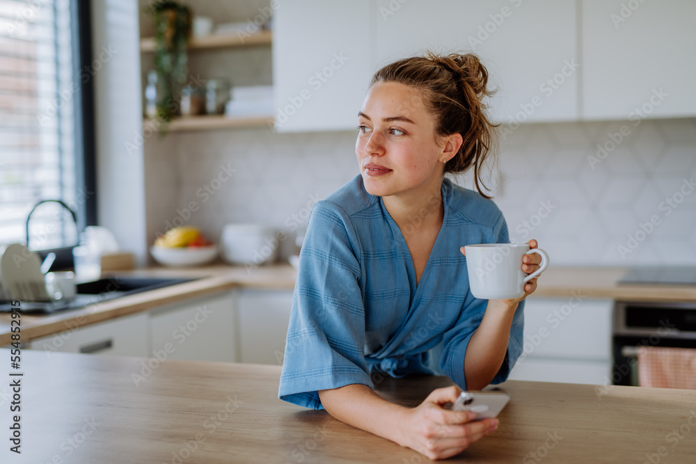 Young woman with smartphone enjoying cup of coffee at morning, in her kitchen. - obrazy, fototapety, plakaty 