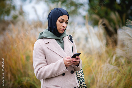 Fotografija Portrait of young muslim woman in coat, standing outdoor in city park, scrolling smartphone