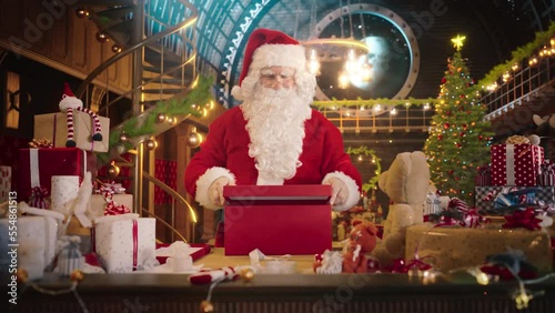 Jolly Santa Claus Works in His Studio Workshop, Wrapping and Packing Christmas Gifts for all the Good Children to be Delivered on the Magical New Year Eve. He Offers the Gifts this Winter Holiday photo