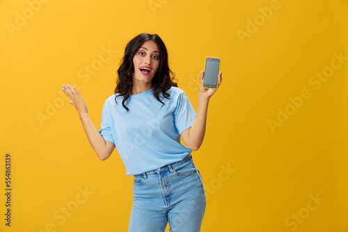 Woman blogger with a phone in her hands in a blue t-shirt and jeans on a yellow background smile signs gestures symbols, online communication and video call, copy space, free background