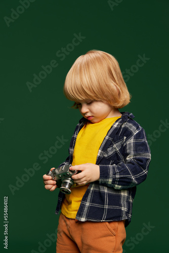 Portrait of little boy, child posing with vintage camera over green studio background. Photographer. Concept of childhood, emotions