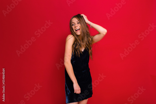 Adorable happy young woman with wonderful smile and long wavy hair wearing stylish festive dress is touching her hair and looking adore while posing over red background 