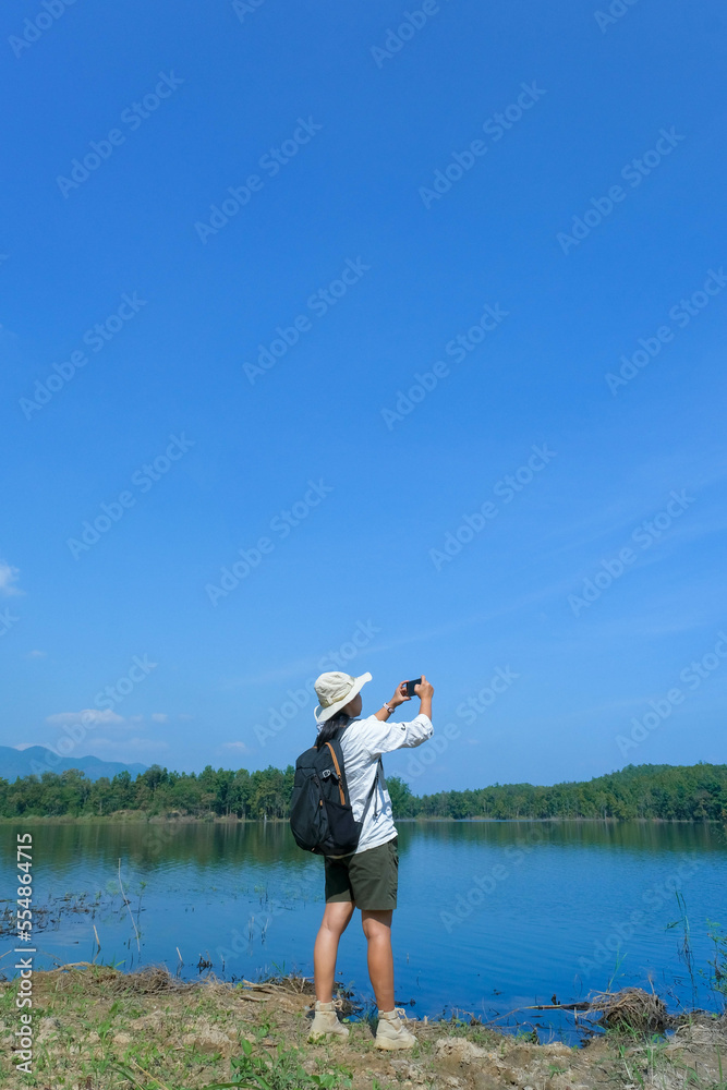 Female ecologist with backpack using mobile phone taking picture of reservoir for data analysis. Female environmental scientist working in field survey.