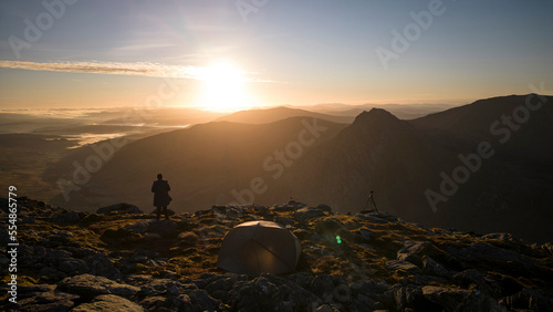 A photographer with camera, tripod and wild camping tent in the mountains