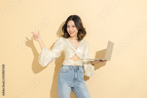 Asian happy woman with perfect body pointing to copy space and holding laptop in studio. Fashionable female model in sexy dress with on beige isolated background. E-commerce, Online shopping Concept.