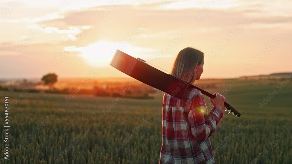 Back view of young female musician walking on field alone at sunset. Pretty, blonde woman looking, smiling, carrying guitar, admiring sunset. Concept of beauty of nature. Back view of young female musician walking on field alone at sunset. Pretty, blonde woman looking, smiling, carrying guitar, admiring sunset. Concept of beauty of nature.