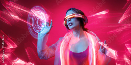 Woman in futuristic costume. Girl in glasses of virtual reality while touching air. Augmented reality game, future technology, AI concept. VR. Neon red light. Viva magenta color of the year 2023. 