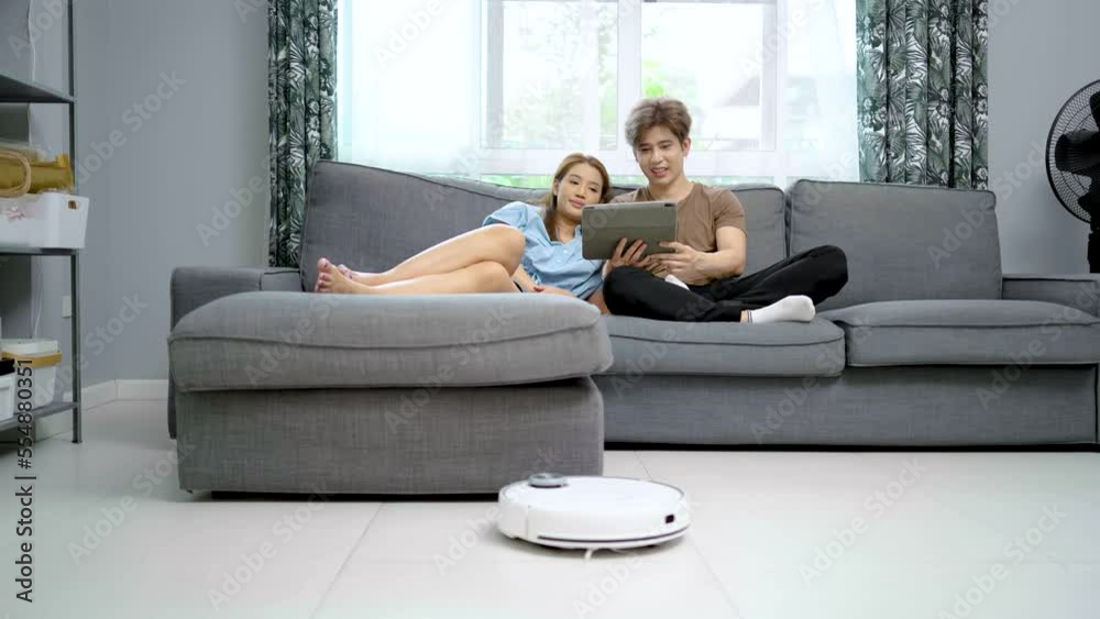 Young couple resting on the couch while robotic vacuum cleaner doing its work at home. Household robots, Smart home concept Young couple resting on the couch while robotic vacuum cleaner doing its work at home. Household robots, Smart home concept