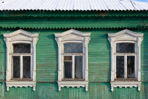 A row of three windows with white carved architraves in an old green rural wooden house. Folk style in architecture