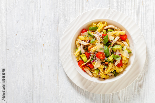 Healthy Chicken Pasta Salad with Avocado, Tomato, and olive oil and vinegar dressing in white bowl on white wood table, horizontal view from above, flat lay﻿, free space