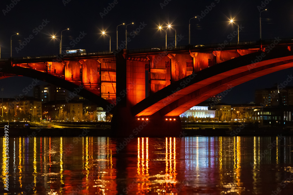Night landscape with red illuminated of Communal Bridge in Krasnoyarsk, Russia. View of the Yenisey River at night. Reflection of lighting lights in the rippled water surface Night landscape with red illuminated of Communal Bridge in Krasnoyarsk, Russia. View of the Yenisey River at night. Reflection of lighting lights in the rippled water surface