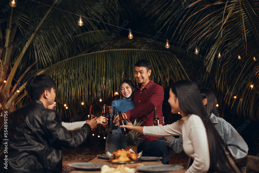 Celebrating anniversary party outdoor of Asian couple with group friends. Happy couple dinner party with friends they clink bottle beer under light in the night. Festival celebrating.