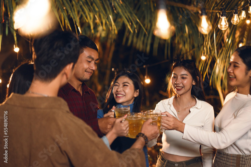 Happy celebrating anniversary party outdoor of Asian couple with group friends. Happy couple dinner party with friends they clink glasses beer under light in the night. Friendship and new year concept