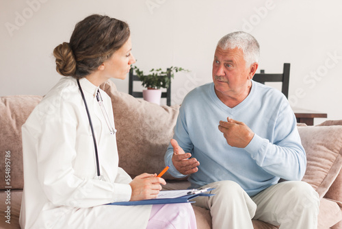 Female professional doctor showing medical test result explaining prescription using clipboard visiting senior elderly old man patient at home sitting on sofa