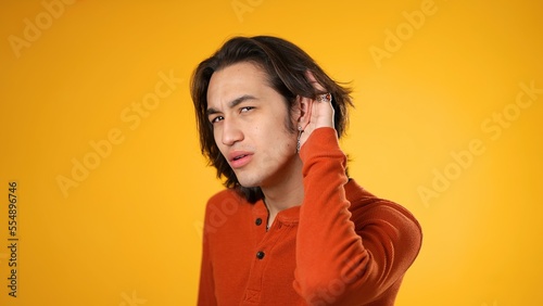 Portrait of Hispanic Latino gender fluid young hipster man 20s puts hand to ear to listen to secret isolated on yellow background studio portrait