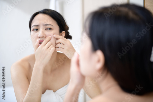Problem skin. Concerned young asian women popping pimple on cheek while standing near mirror in bathroom. young asian women with acne