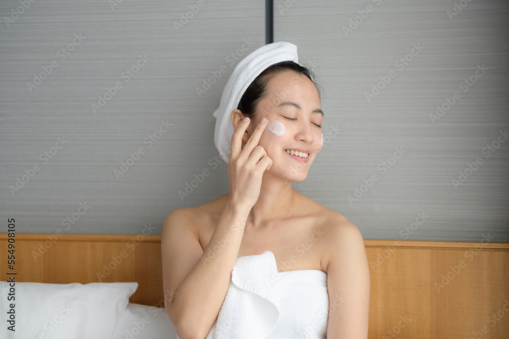 Happy young Asian woman applying face lotions while wearing a towel and touching her face. Daily makeup and skincare Happy young Asian woman applying face lotions while wearing a towel and touching her face. Daily makeup and skincare