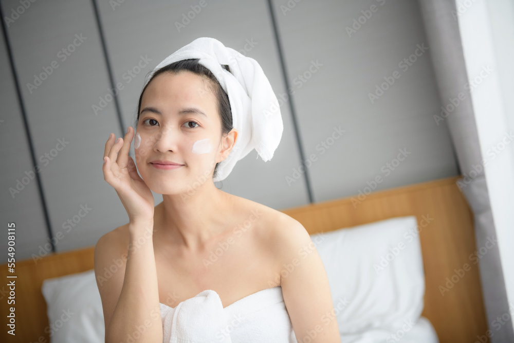 Happy young Asian woman applying face lotions while wearing a towel and touching her face. Daily makeup and skincare Happy young Asian woman applying face lotions while wearing a towel and touching her face. Daily makeup and skincare