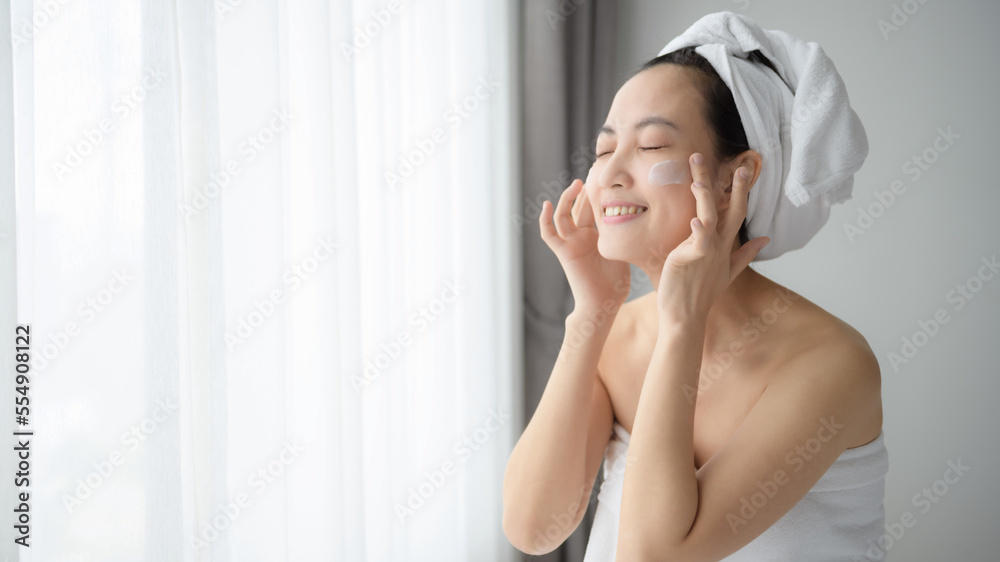 Happy young Asian woman applying face lotions while wearing a towel and touching her face. Daily makeup and skincare Happy young Asian woman applying face lotions while wearing a towel and touching her face. Daily makeup and skincare